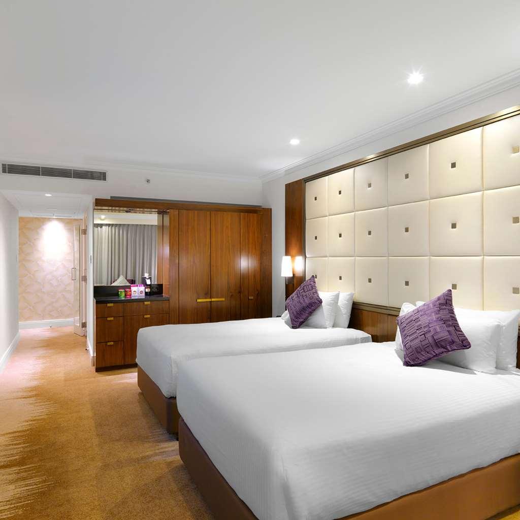 Amora Hotel Jamison Sydney Rom bilde The photo shows a modern hotel room featuring two twin beds with white bedding and purple decorative pillows. The walls are designed with soft beige panels, creating a luxurious ambiance. There’s wooden furniture, including a cabinet against one wall